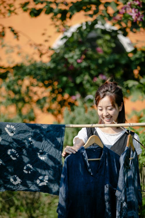 Free Cheerful Asian female near rope with painted textile and garments on hangers standing on sunny street with green plants and building on blurred background Stock Photo