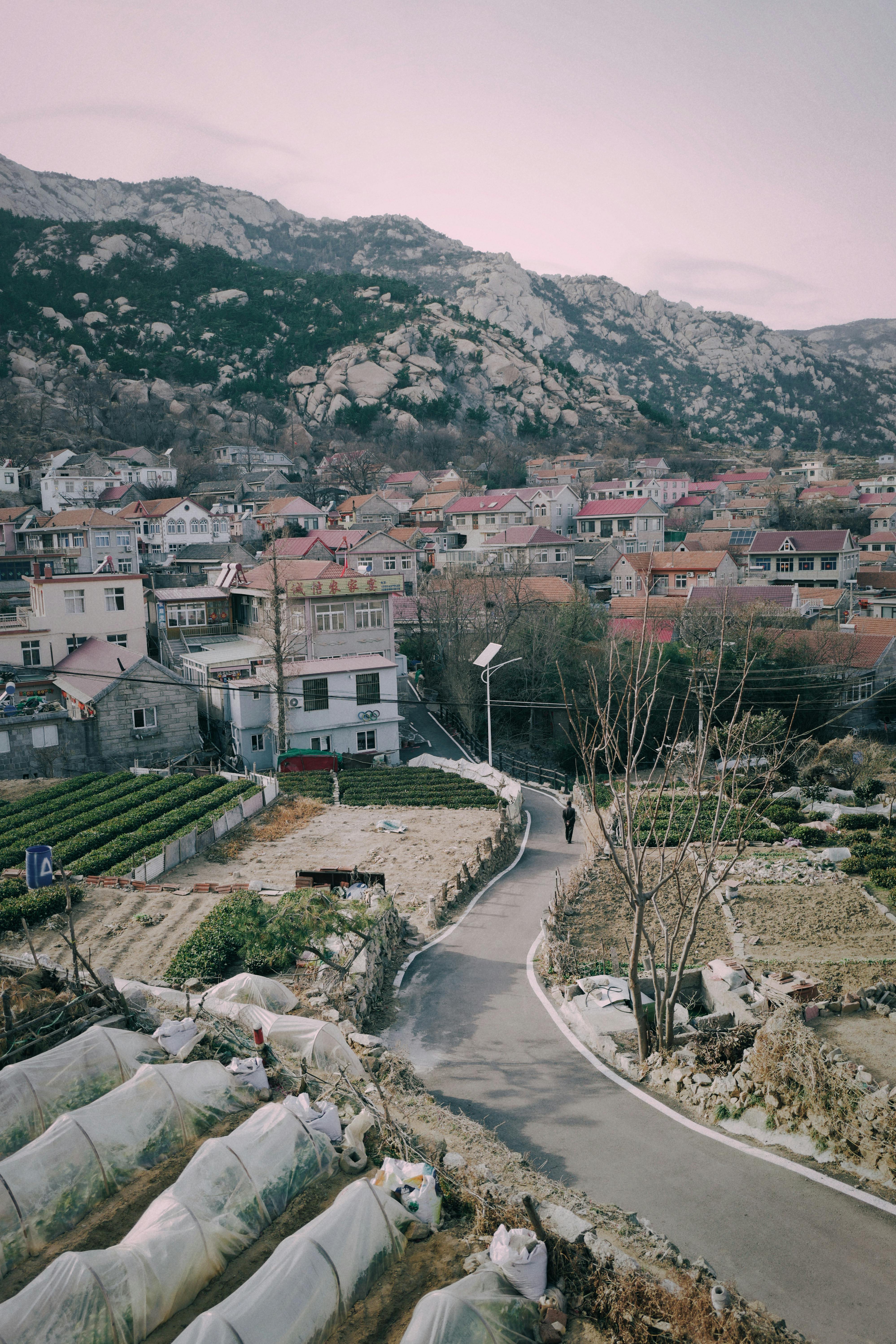 Scenic view of a rural village with a winding road and fields under mountainous backdrop.