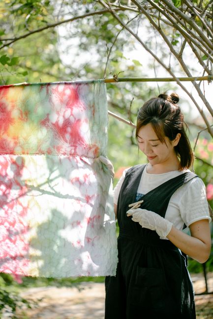 Positive Asian female in uniform standing near hanging colorful fabric dyed using shibori technique standing on sunny street with green trees