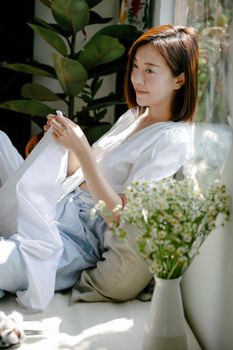 Smiling young ethnic lady in casual clothes sitting on sofa while sewing white fabric near vase with flowers in living room in sunny day while looking away