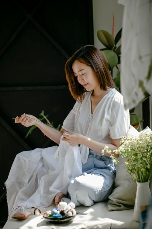 Free Young Asian lady in casual outfit sewing white fabric while sitting on couch with pillow and bowl with threads near vase with flowers in living room in sunny day Stock Photo