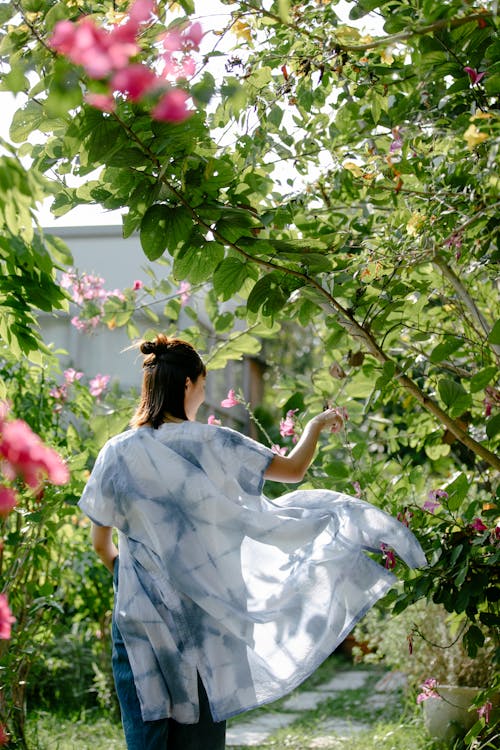 Back view of anonymous female artisan in clothes with indigo ornament among blooming plants in sunlight