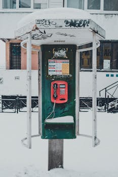 Shabby public pay phone booth placed on street covered with snow against residential building in city on cold winter day