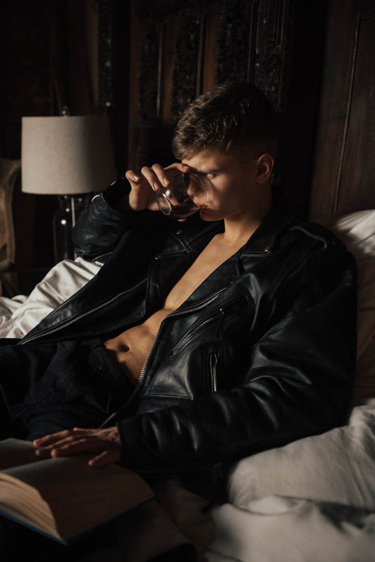 A Man In Black Leather Jacket Sitting On Bed While Drinking