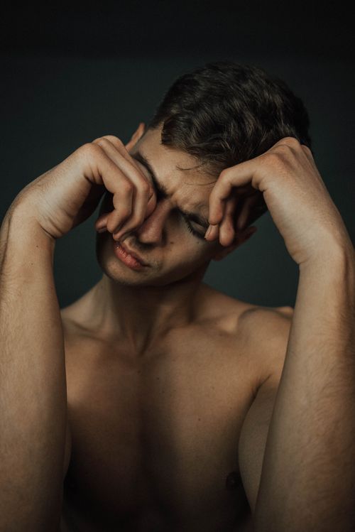 Free A shirtless young man rubs his eyes, conveying emotion and vulnerability in a dark setting. Stock Photo