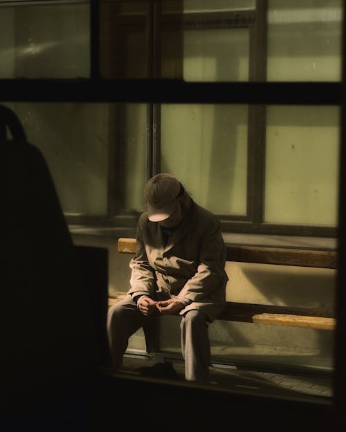 Through glass of aged male in outerwear sitting on bench and waiting for transport