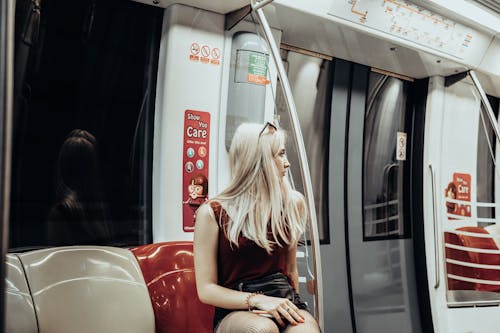 Young female passenger with long blond hair in stylish outfit sitting on seat and looking away while commuting by modern train