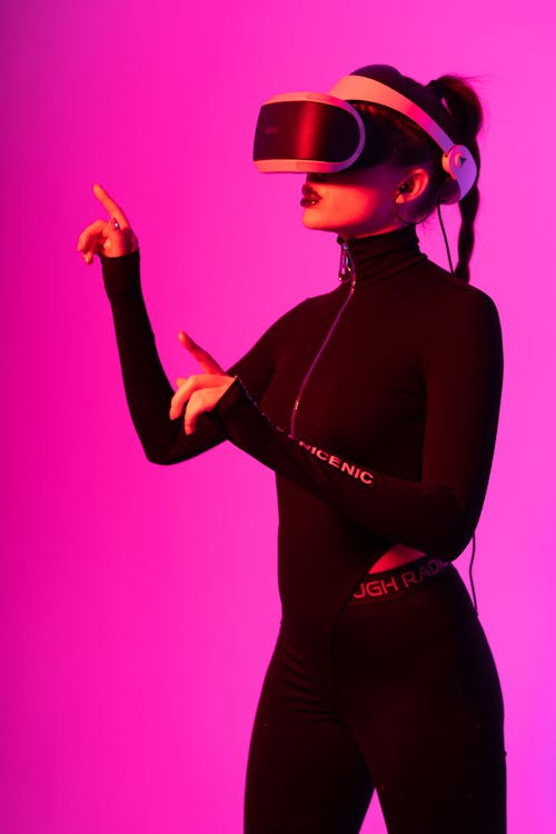 Young fashionable female using VR headset while entertaining and exploring cyberspace on pink background