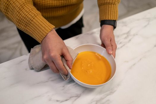 Person serving creamy pumpkin soup in a ceramic bowl on a marble surface.