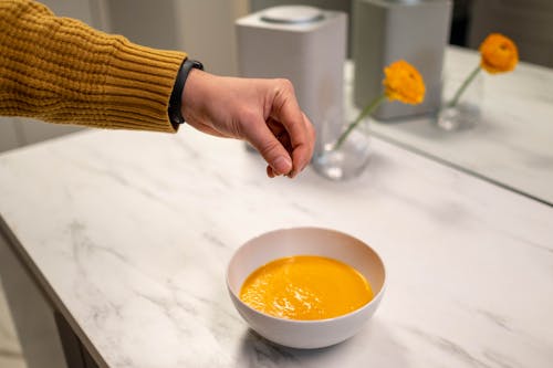 Free A hand sprinkles seasoning on freshly made pumpkin soup in a modern kitchen setting. Stock Photo