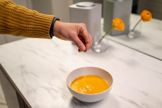 A hand sprinkles seasoning on freshly made pumpkin soup in a modern kitchen setting.