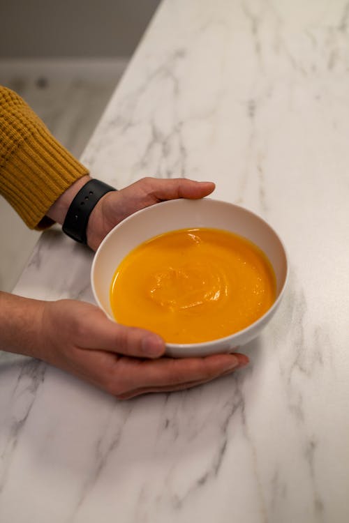 Hands Holding a White Ceramic Bowl with Pumpkin Soup
