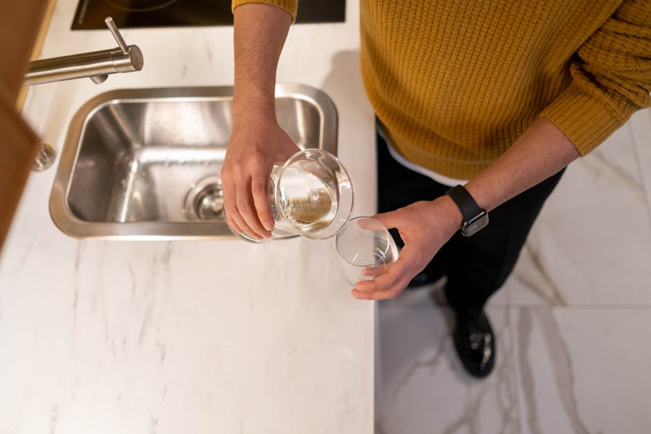 A person wearing a yellow sweater pours water into a glass at a kitchen sink.
