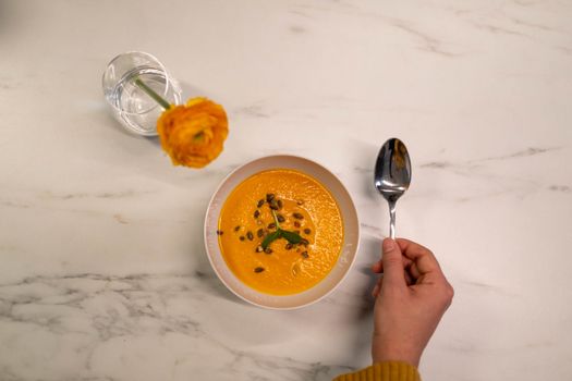 A top-down view of creamy pumpkin soup garnished with seeds and herbs, accompanied by a bright yellow flower in a glass vase.