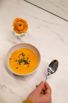 Top view of pumpkin soup garnished with seeds beside a vibrant orange flower.