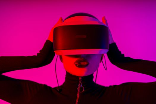 Astonished young female using virtual reality goggles while experiencing invisible reality on pink background