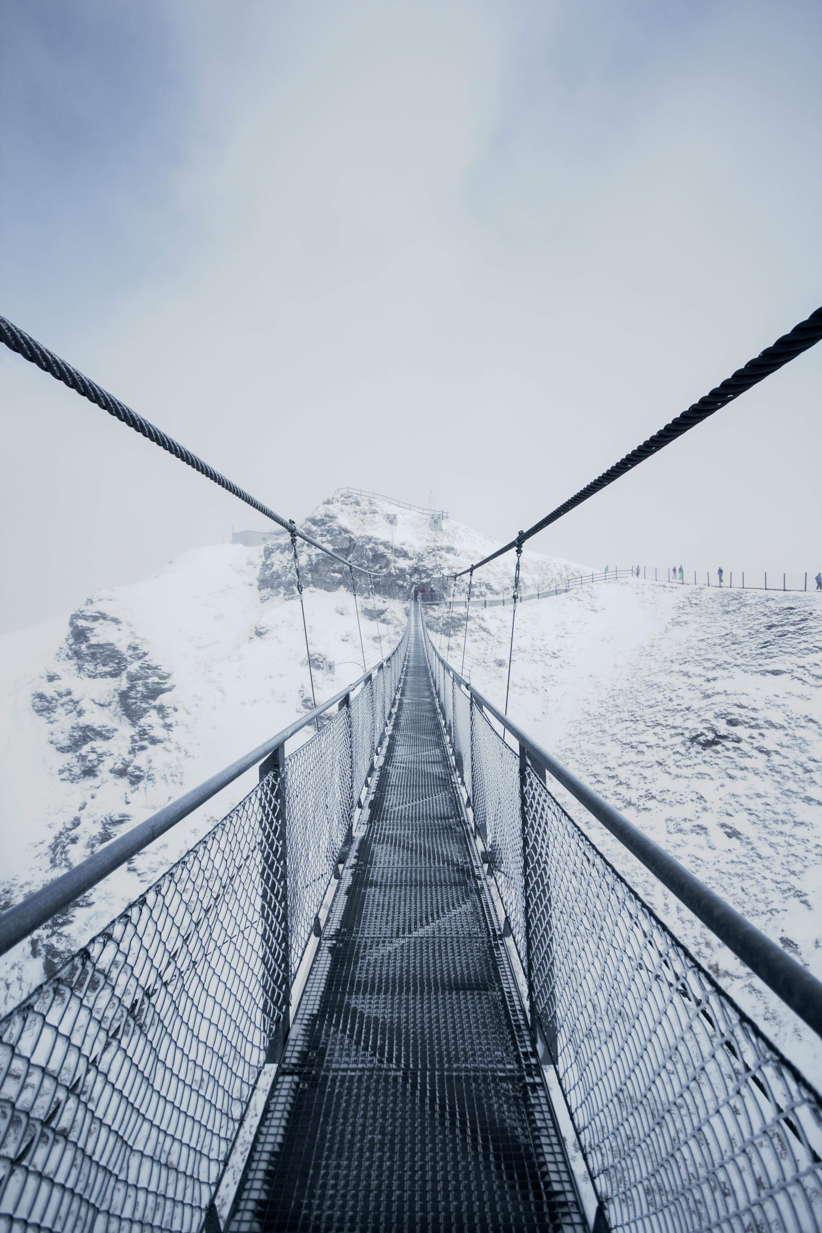 A dramatic view of a metal suspension bridge over a snowy, mountainous terrain, showcasing winter's serene beauty.