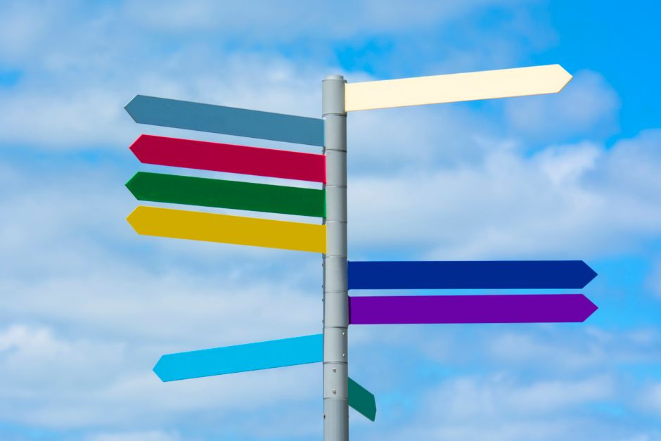 A grey metal post carrying nine blank arrow-shaped direction signs in cream, grey-blue, red, green, yellow, dark blue, purple, light blue and teal, pointing left and right against a blue sky with white cloud