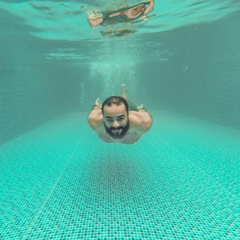 A joyful bearded man swimming underwater in a clear pool with a tiled floor.