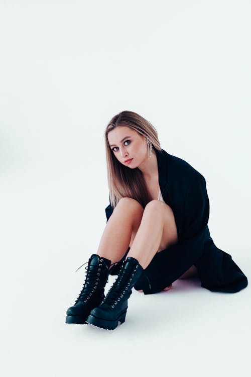 Full body of young serious female in trendy outfit sitting on floor and looking at camera in light studio on white background