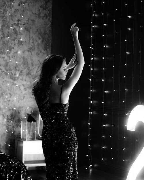 Black and white back view of confident young female in shiny dress dancing with raised hands and closed eyes near illuminated garlands in light room