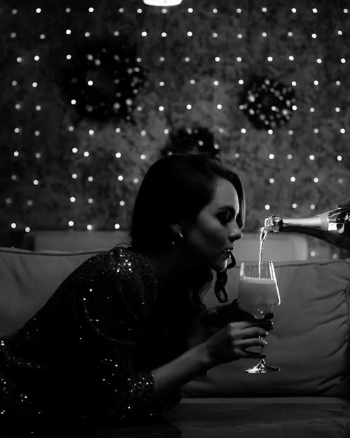 Black and white of young thoughtful woman in shiny dress lying on bed with closed eyes while person pouring alcohol from bottle in glass near glowing garlands in hotel room