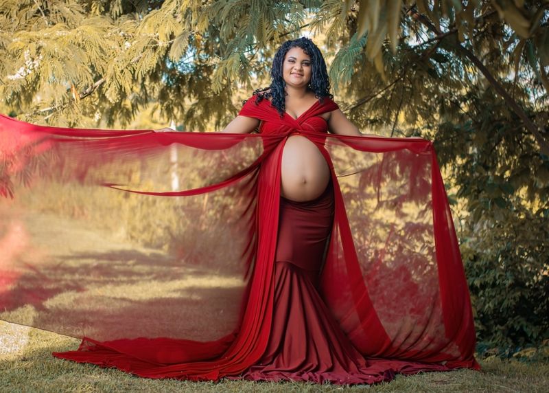 Pregnant woman in a flowing red dress posing confidently outdoors, showcasing her baby bump.