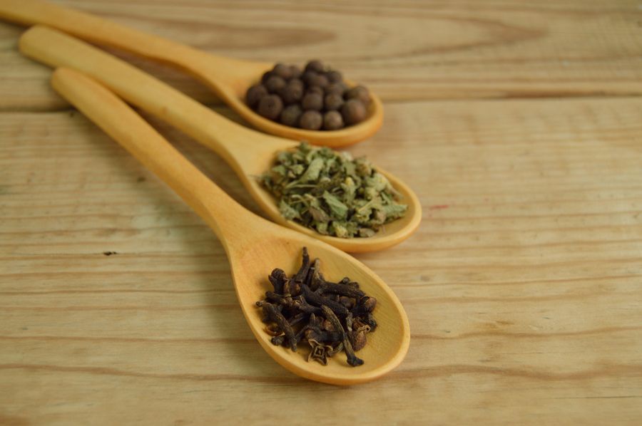 Three wooden spoons holding allspice berries, dried herbs and whole cloves on a pale wood table