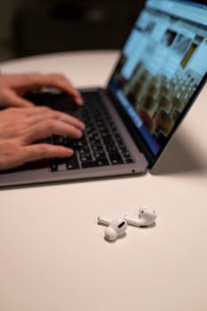 Hands typing on laptop with wireless earbuds on table, indoors.