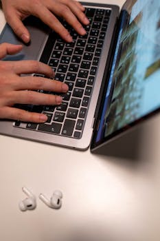 Close-up of hands typing on a laptop keyboard with AirPods placed beside it on a desk.