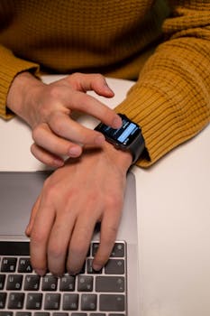 Close-up of a man interacting with a smartwatch while working on a laptop in an indoor setting.