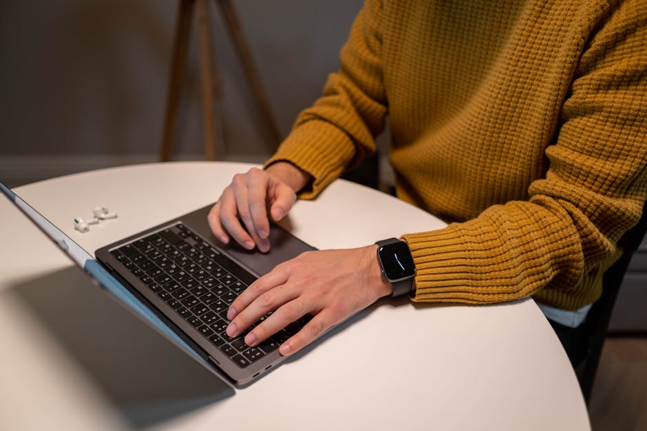 Apple Watch Ultra 2 vs Garmin Fenix 8: Battery Life for Multi-Day Hiking (2024) A man in a yellow sweater uses a laptop and smartwatch at a table. AirPods are visible beside him.