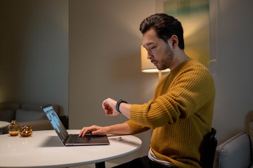 Free Man in Brown Sweater Looking at Time on His Smartwatch while Using Laptop Stock Photo