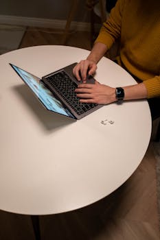 Man typing on laptop with smartwatch and earbuds on a round table indoors.