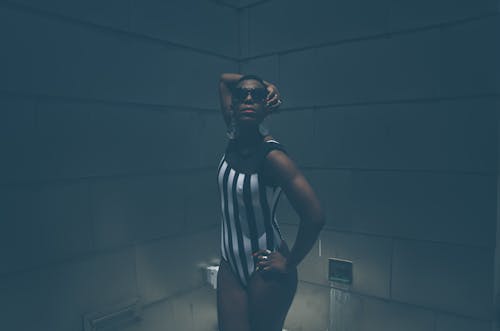 Eccentric young black transgender male with short hair in stylish bodysuit and sunglasses standing in dark studio with hand on waist