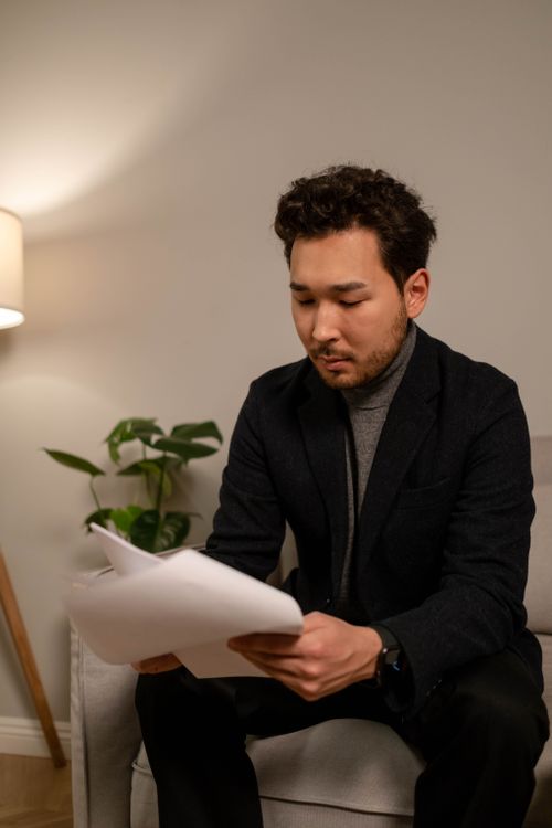 Free Asian man in a suit reading documents indoors, creating a focused and professional atmosphere. Stock Photo