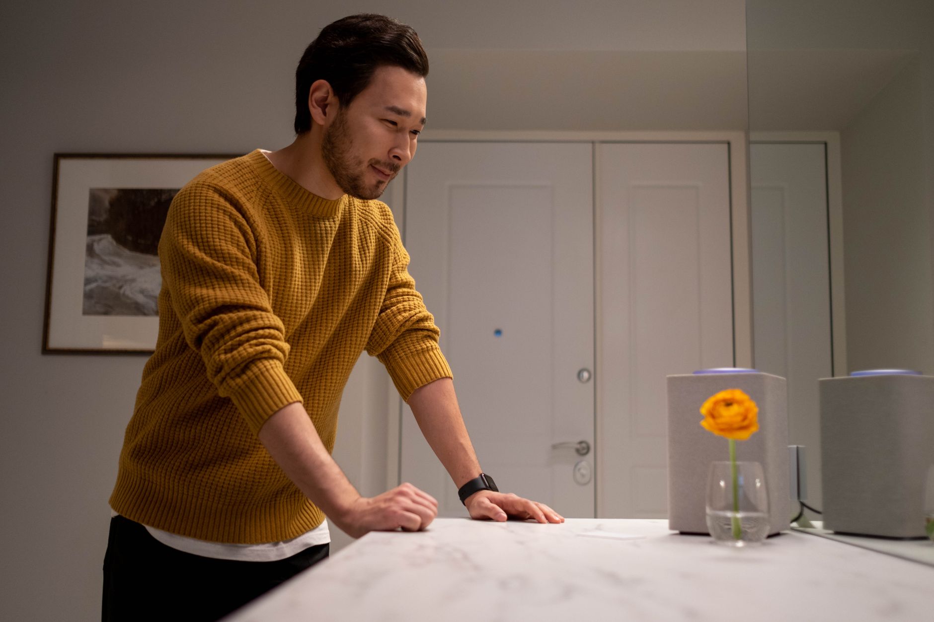 A man in a mustard sweater smiling at a smart speaker in a modern interior setting.