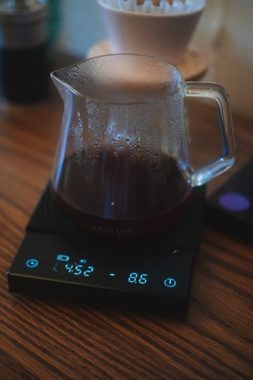 Modern electronic drip coffee scale with glass pot with hot beverage placed on wooden table with cup on blurred background