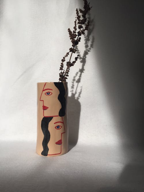 Minimalistic beige vase with dried twig of plant placed near while wall and illuminated by sunlight