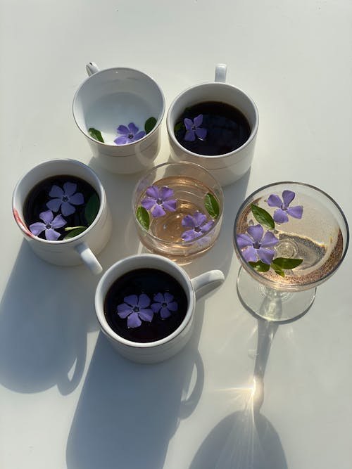 Free From above of cups of tea and water and glasses with drinks decorated by purple flowers of Vinca minor against light table Stock Photo