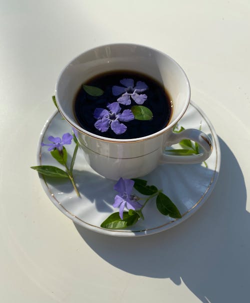 From above of ceramic saucer with cup of drink decorated with blue periwinkle flowers and green leaves on white surface