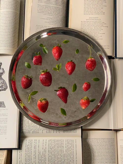 From above of metal round tray with sliced red strawberries and green leaves placed on opened books