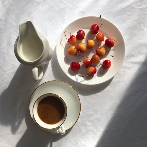 Top view of ripe sweet cherries on plate placed on white cloth near mug of hot coffee and saucer with milk