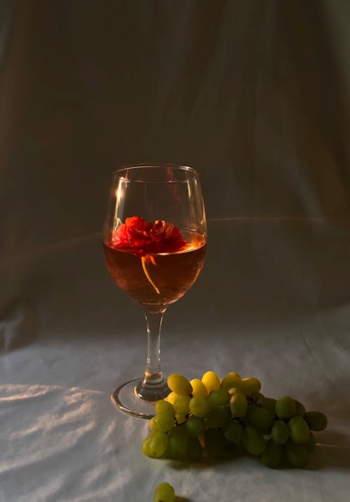 Transparent wineglass with alcoholic red drink and small flower placed on white creased fabric with fresh green grapes in studio