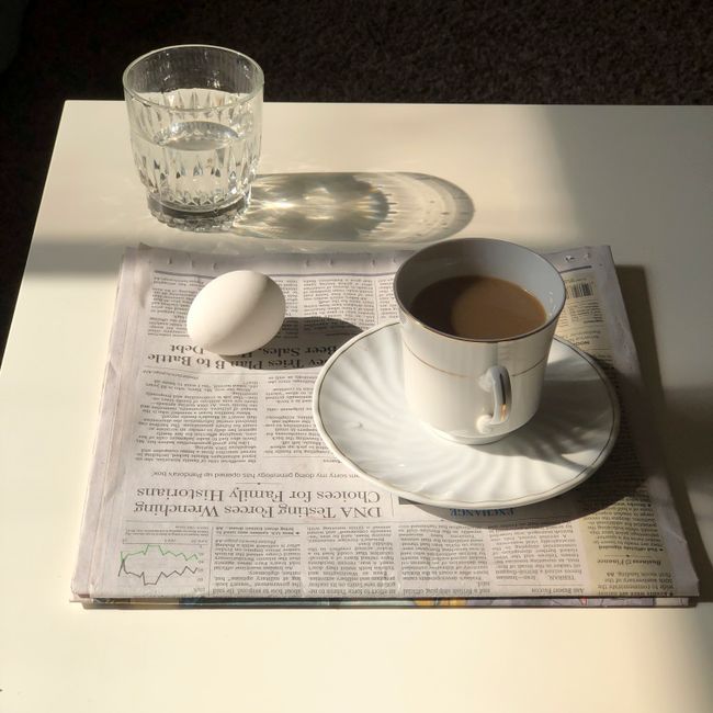 Composition of freshly brewed coffee with milk in white cup placed near boiled egg on morning newspaper