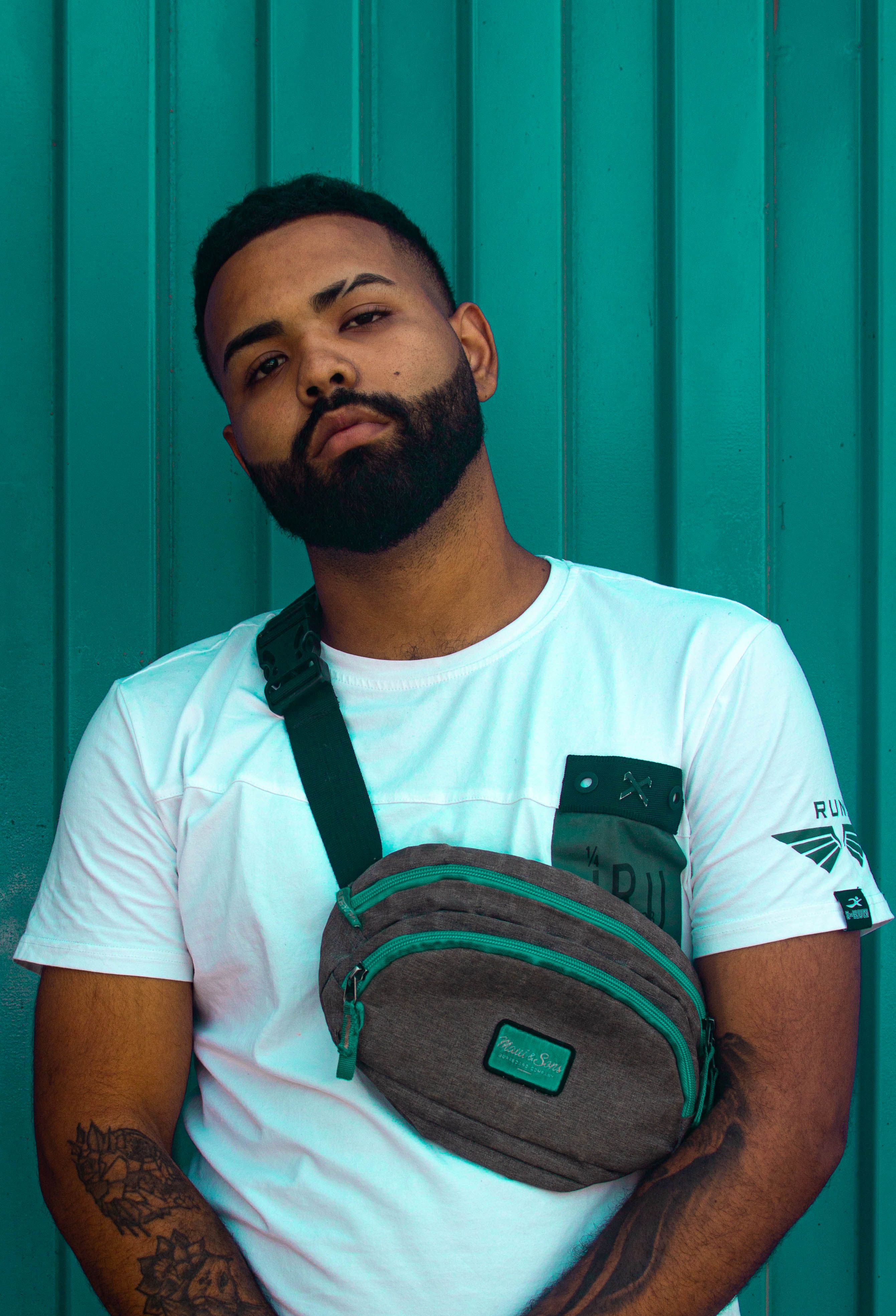 A young man with facial hair poses stylishly with a sling bag against a teal background.