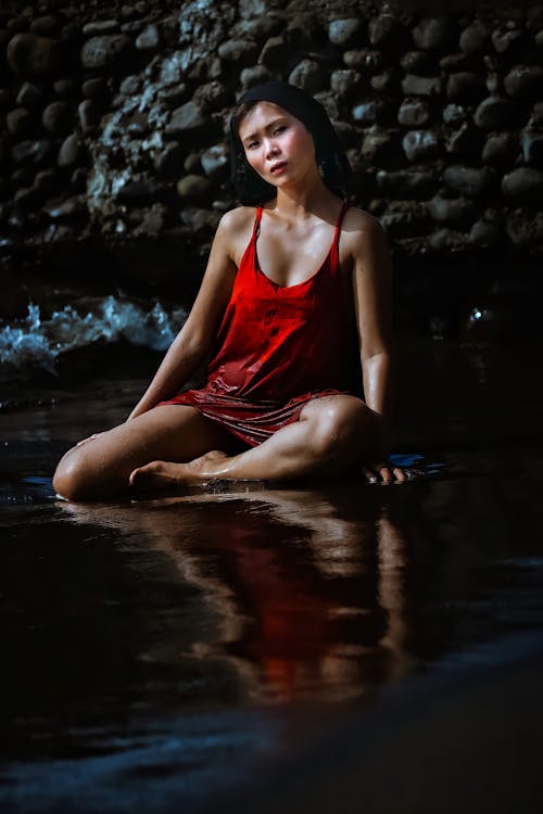 Ethnic female in dress sitting in water near stones