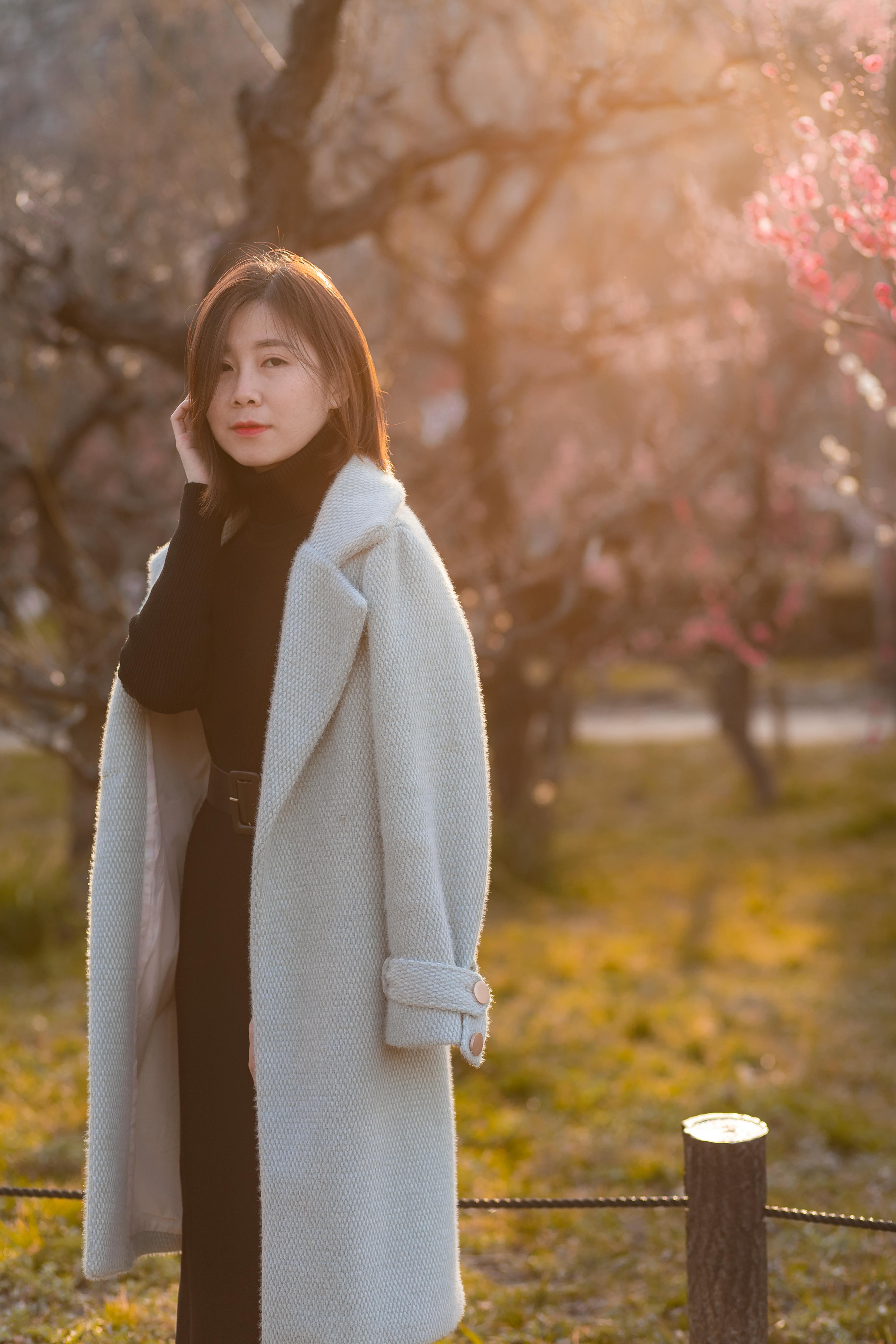 A stylish woman in a coat poses gracefully against a sunny park backdrop with blossoming trees.