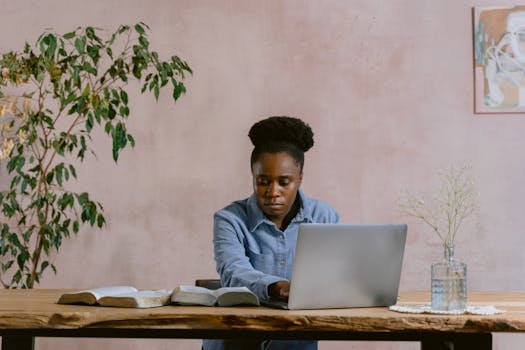 A woman sits at a table with a laptop and open books, reflecting concentration and learning.