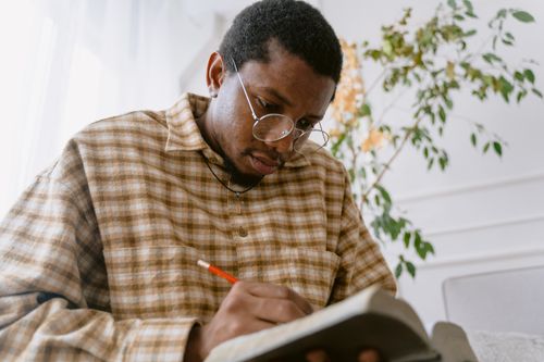 Free African American man wearing eyeglasses writing in a notebook indoors with natural light. Stock Photo
